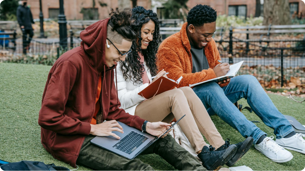 Three students sitting on a grass lawn, studying with a laptop, book, notebook.